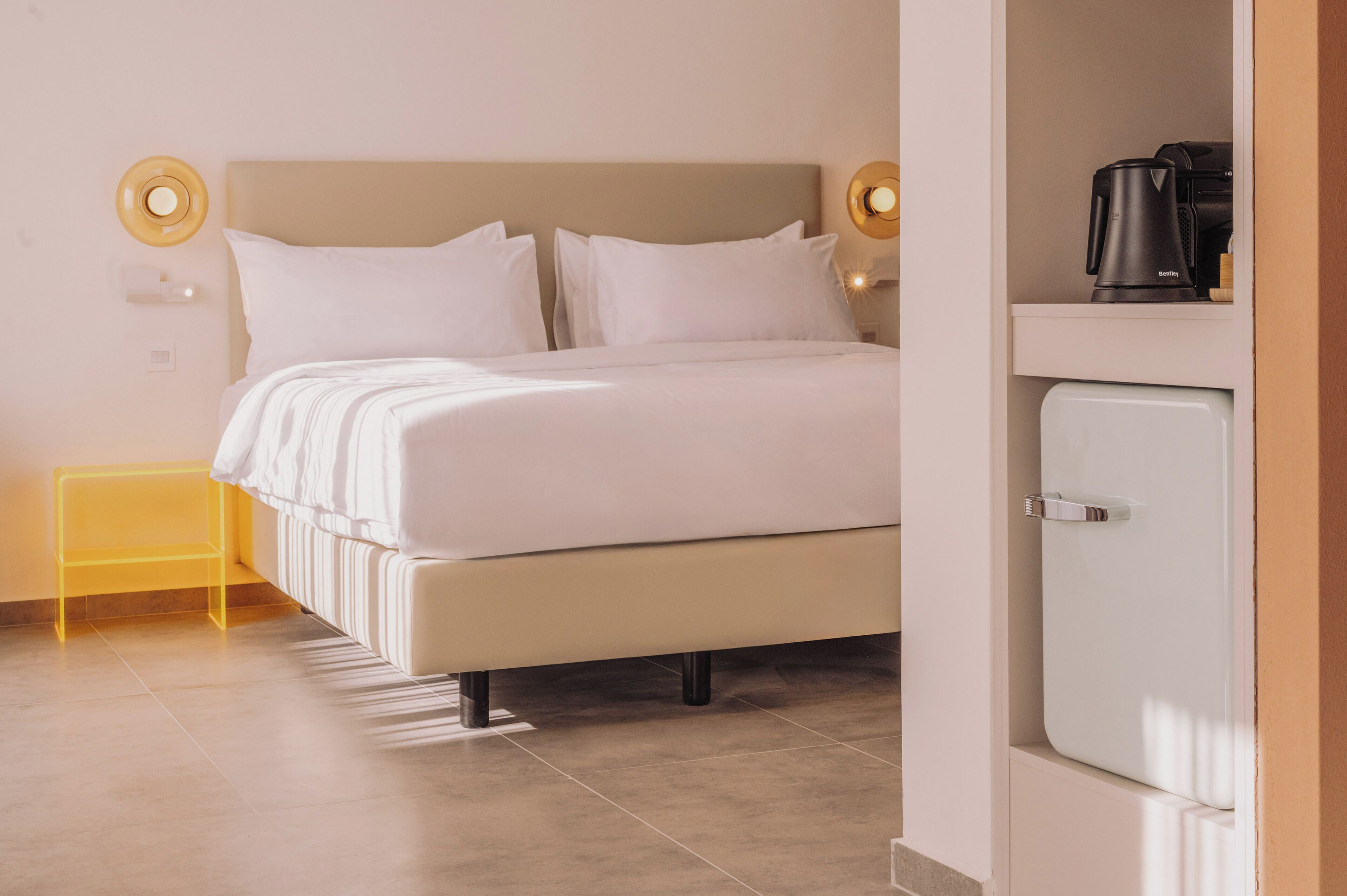 A neatly made bed with white bedding and pillows, a small bedside table with a yellow lamp, and a white cabinet with a coffee maker.