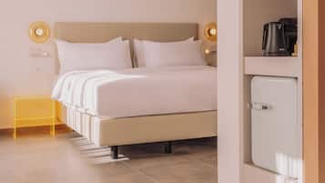 Una cama bien hecha con ropa de cama y almohadas blancas, una mesita de noche con una lámpara amarilla y un armario blanco con cafetera.