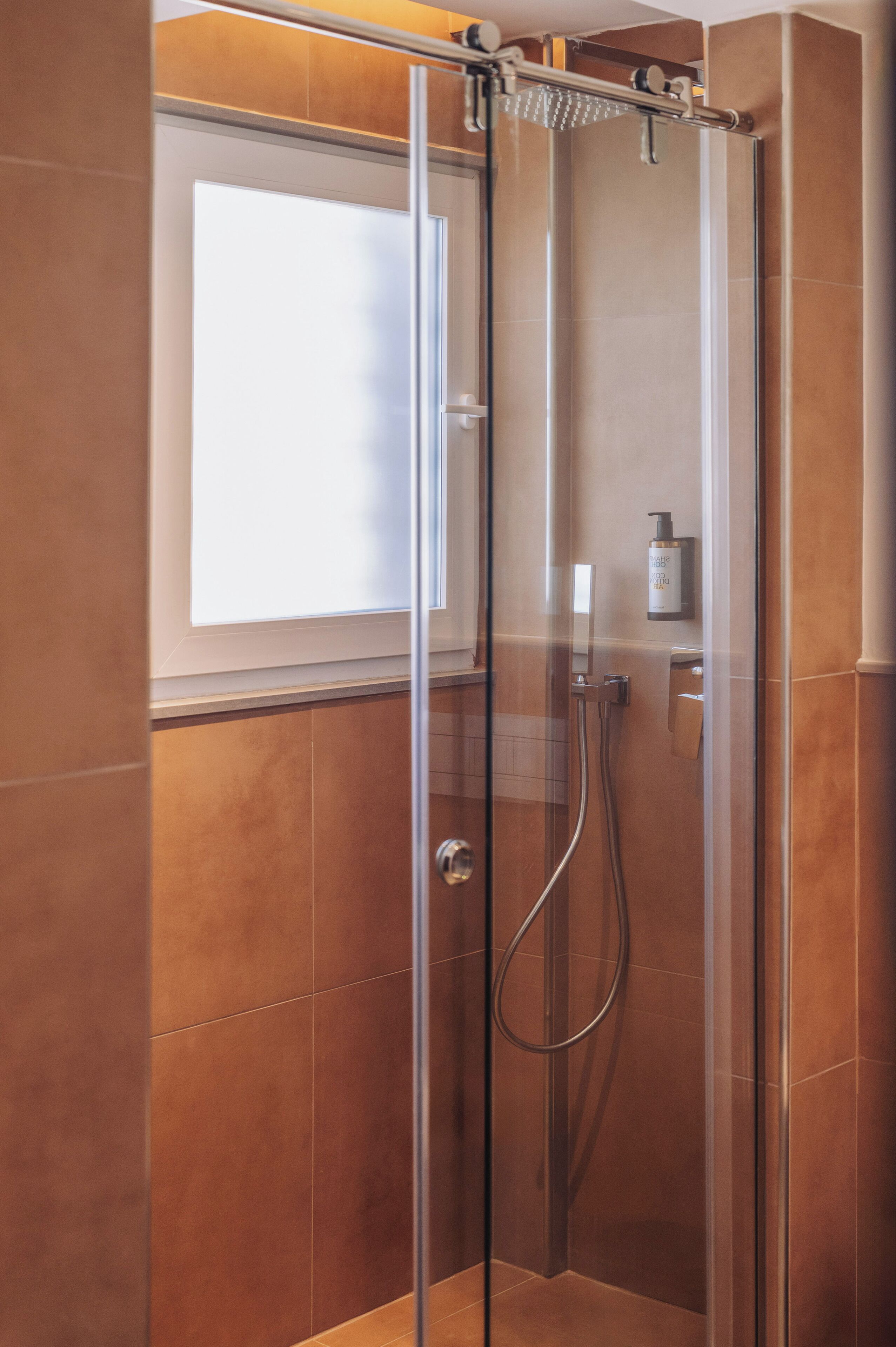 A glass-enclosed shower with a handheld showerhead, a bottle of shampoo, and a towel rack.