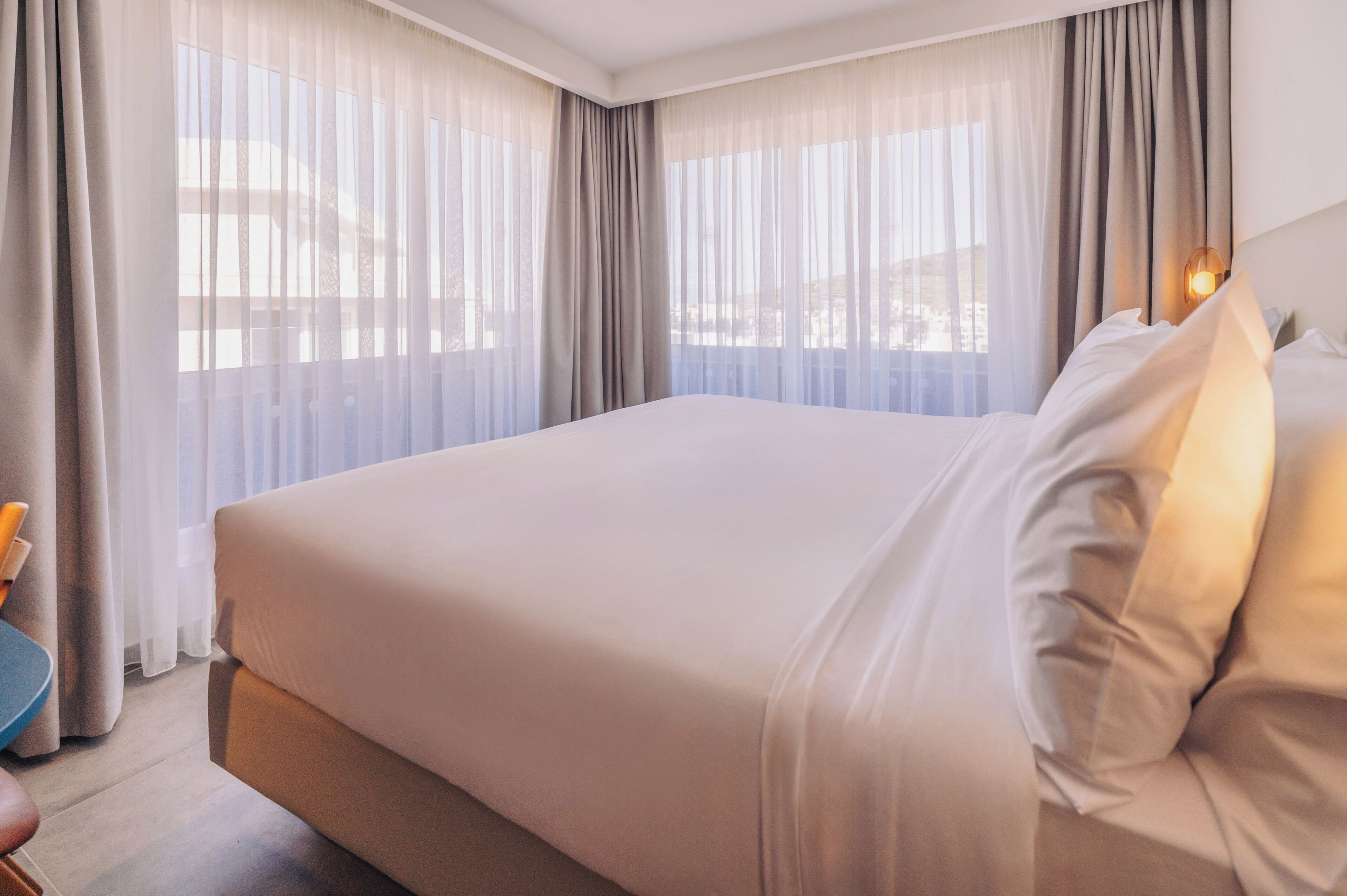 A hotel room with a large bed, sheer curtains, and a nightstand with a lamp.