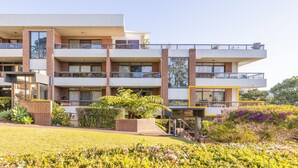 A modern multi-story building with balconies, a landscaped garden, and a clear sky.