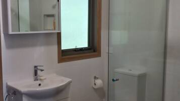 A bathroom with a white sink, mirror, and toilet. There is a window above the sink and a glass shower enclosure.