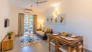 A compact living space with a sofa, dining table, and a TV mounted on the wall. - Palm Retreat 1BHK Pool & Field Views (Calangute)
