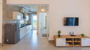 A modern kitchen with a grey refrigerator, white cabinets, and a tiled floor. A wooden cabinet with a plant and a TV is visible. - Palm Retreat 1BHK Pool & Field Views (Calangute)
