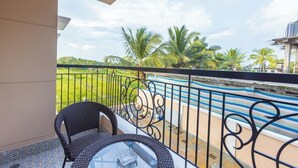 A balcony with a wicker chair and table, overlooking a pool with palm trees and a grassy area. - Palm Retreat 1BHK Pool & Field Views (Calangute)