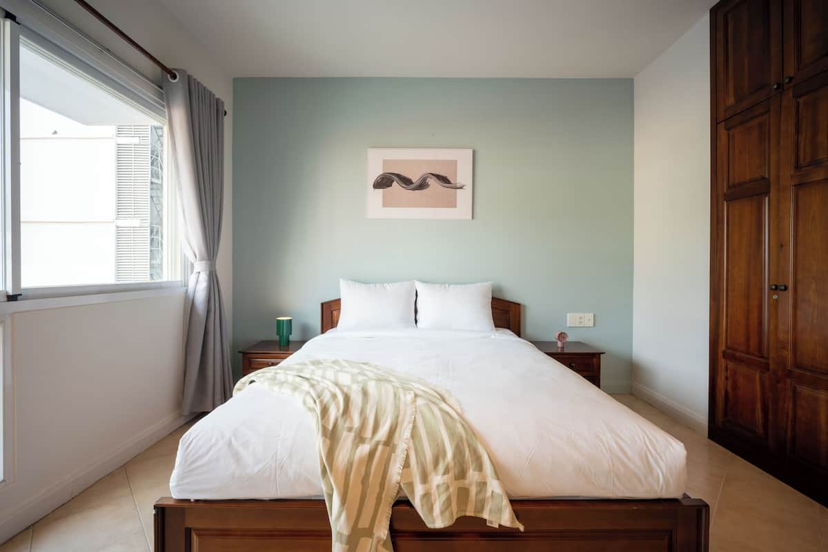 Un lit bien fait, avec une couette blanche et un plaid à rayures, une œuvre d’art encadrée au mur et une armoire en bois.