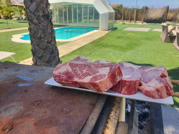 A grill with raw meat, a pool, and a greenhouse in the background.