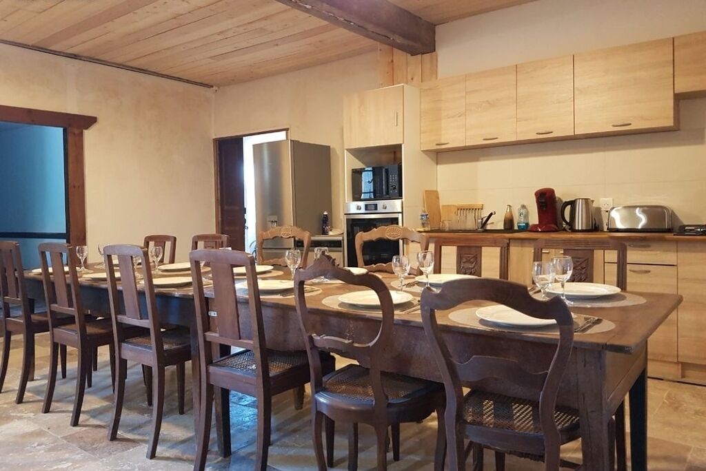 A dining area with a long wooden table set for six, surrounded by six chairs. A kitchen with wooden cabinets and appliances is visible in the background.