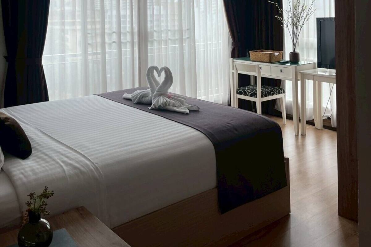 A hotel room with a large bed, a desk, a chair, an air conditioning unit, and a view through sheer curtains.