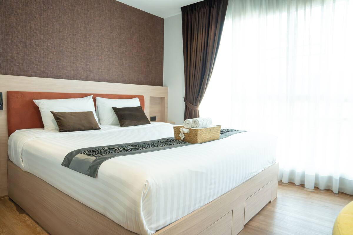 A neatly made bed with a headboard, a bedside basket with towels, and a window with curtains.