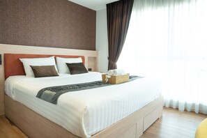 A neatly made bed with a headboard, a bedside basket with towels, and a window with curtains. - Shine Thonglor (Bangkok)