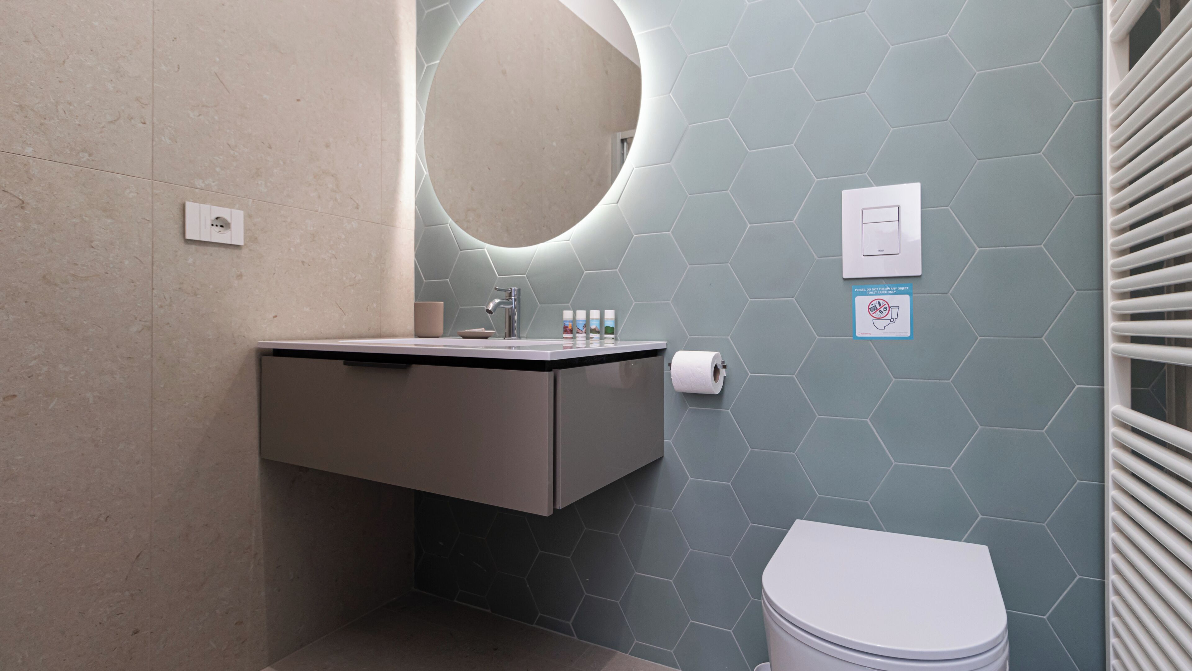 A modern bathroom with a hexagonal tile wall, a round mirror, a sink with a black countertop, a toilet, and a towel rack.