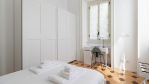 A bedroom with a bed, a desk, and a chair. There are white walls and a window with blinds.
