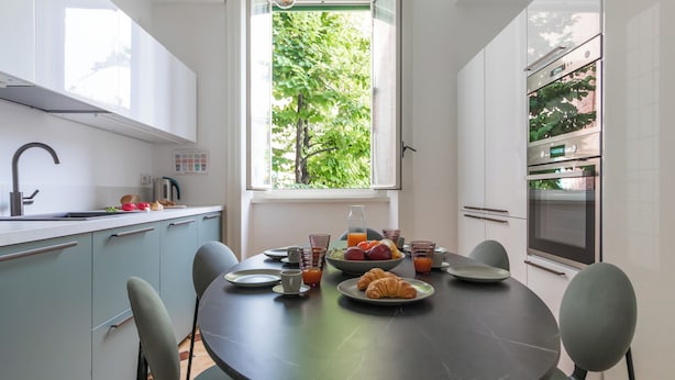 A modern kitchen with a dining table set for breakfast, a window with a view of trees, and built-in appliances.