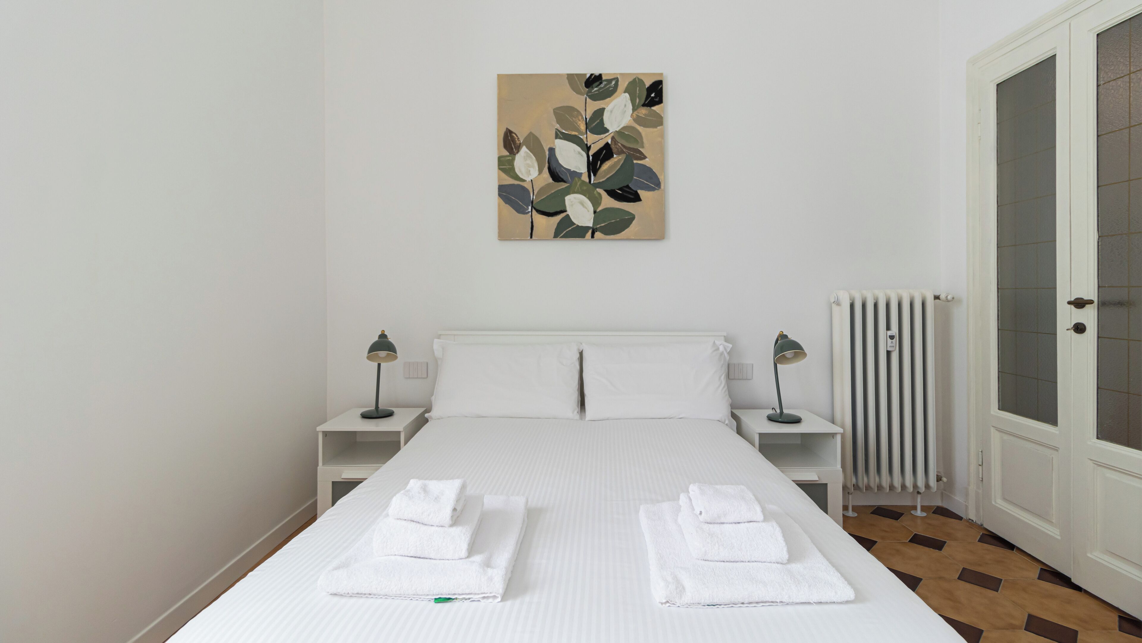 Un lit bien fait, avec du linge de lit et des serviettes blanches, une table de chevet blanche avec une lampe, un radiateur et une porte dotée de panneaux en verre dépoli.