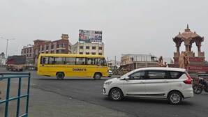 Un autobus jaune portant l’inscription « CHANAKYA » est garé dans une rue de la ville, avec des bâtiments et un monument en arrière-plan.