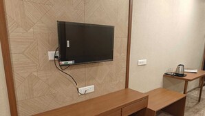 Une pièce avec un téléviseur fixé au mur, un mur orné d’un motif, un banc en bois, une petite table avec une bouilloire et une prise de courant.