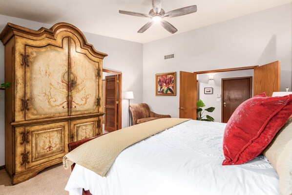 A bedroom with a large wooden wardrobe, a bed with a red pillow, a ceiling fan, and a door leading to another room.