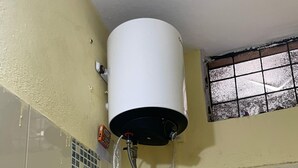 Une salle de bain avec un chauffe-eau blanc fixé au mur, un sol carrelé et une fenêtre avec des barreaux.