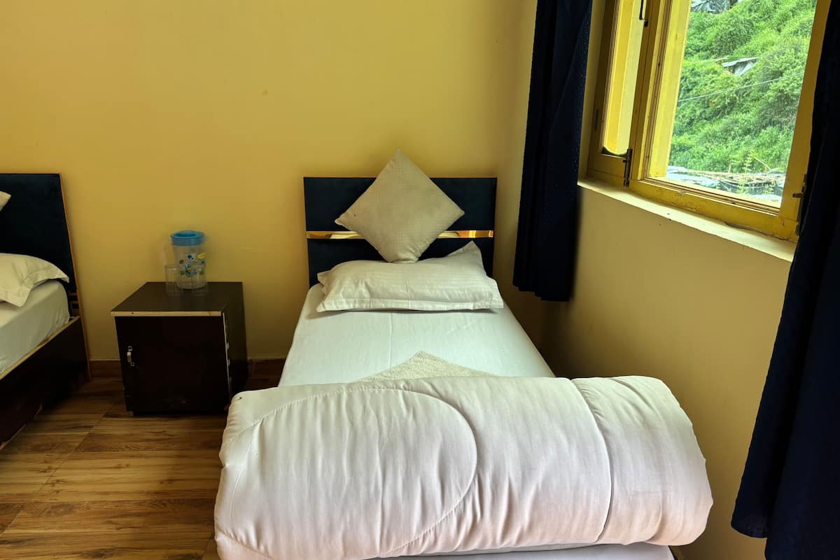 Una cama individual con ropa de cama blanca, una mesita de noche con una botella de agua y una ventana con cortinas.