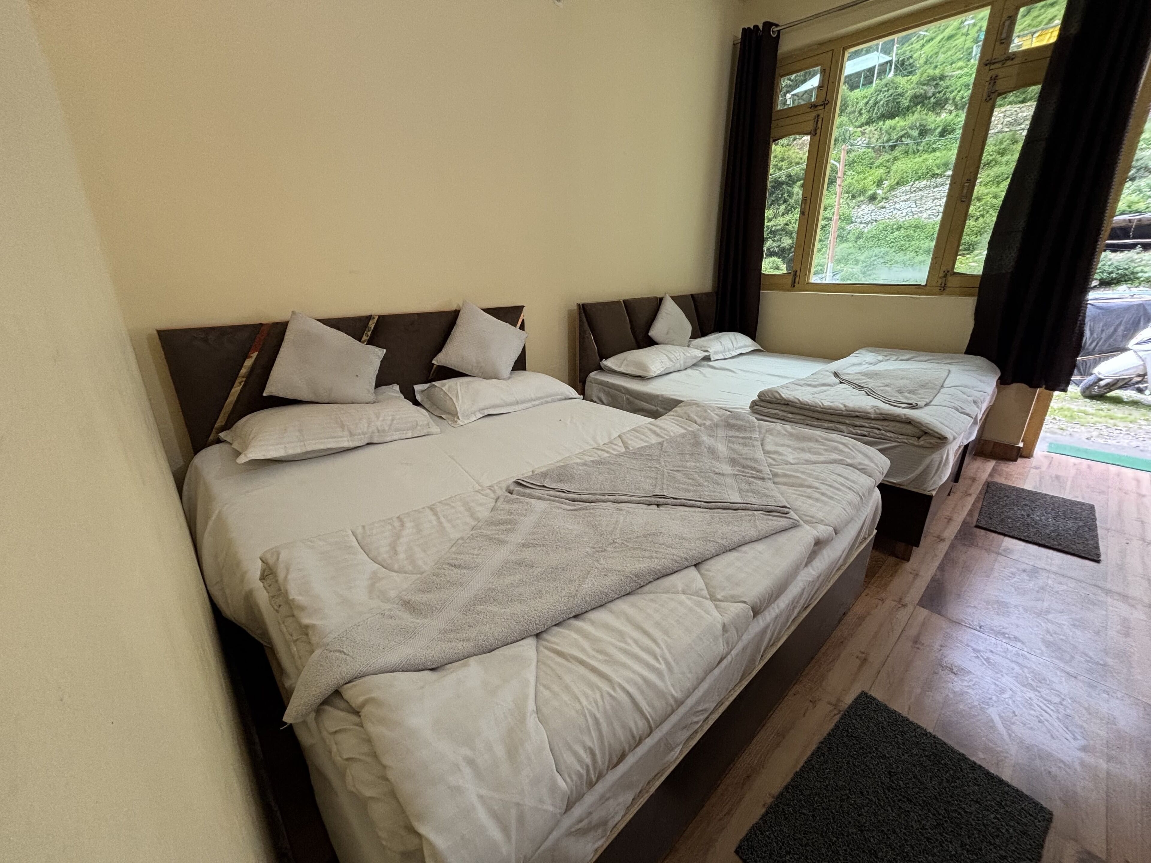 A room with two beds, each with a headboard and pillows. There is a window with curtains, and a door leading outside.
