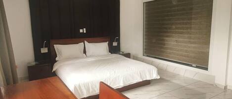 Una camera d'albergo con un letto grande, un comodino in legno, un orologio da parete rotondo e un'ampia finestra con tende.