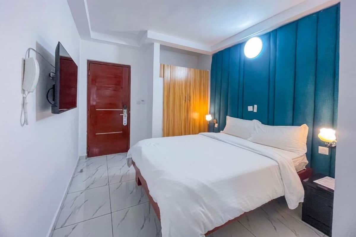 A hotel room with a bed, a wooden door, a wall-mounted phone, and a nightstand with a lamp.