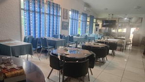 Un comedor con mesas y sillas redondas, cortinas azules y una cocina al fondo.