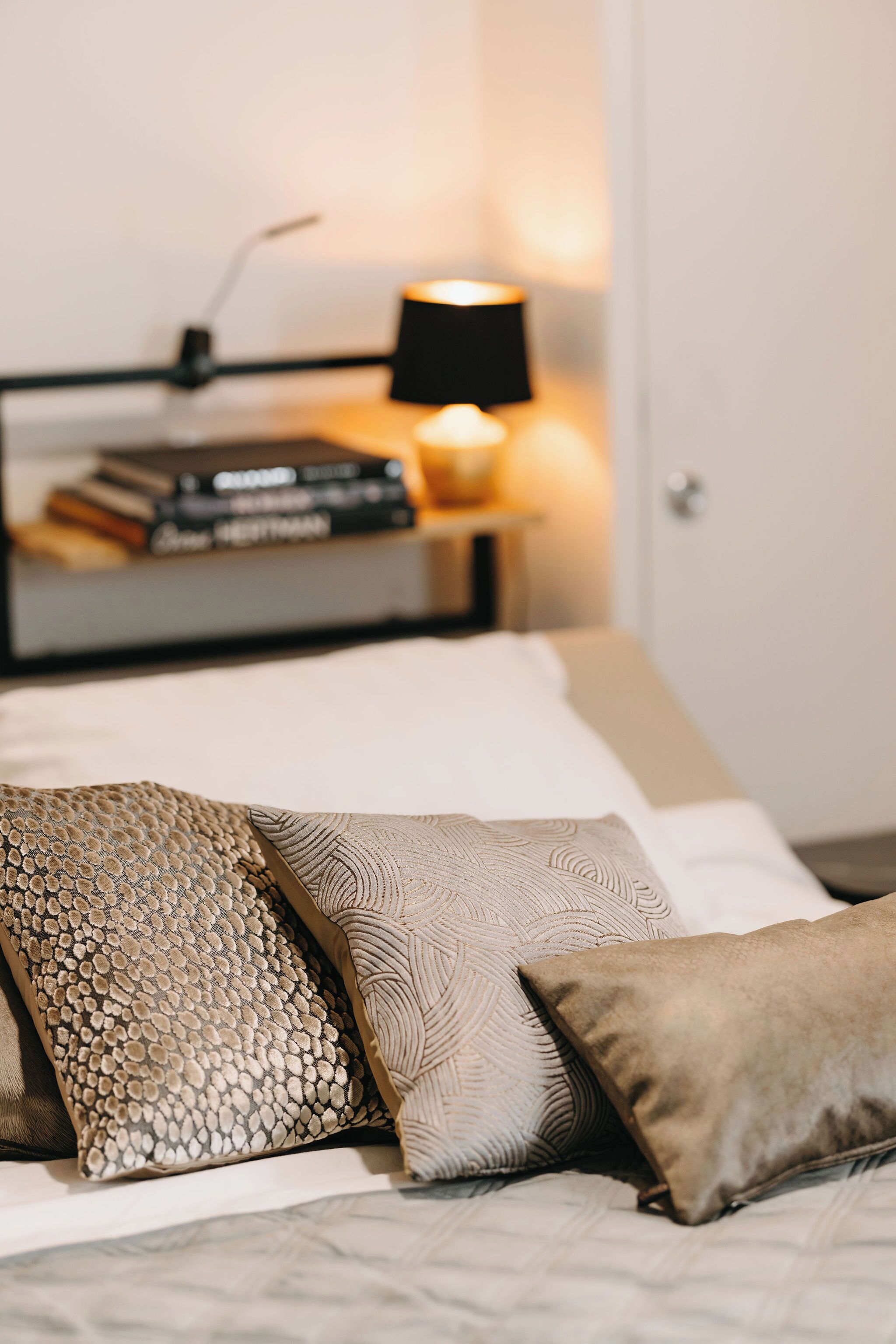Een net opgemaakt bed met gekleurde kussens en een nachtkastje met een lamp en boeken.
