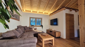 A cozy living room with a sectional sofa, a wooden coffee table, a television, and a window with a view of trees.