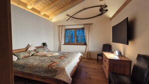 A bedroom with a wooden ceiling, a bed with patterned bedding, a window with curtains, a desk with a chair, and a flat-screen TV mounted on the wall.