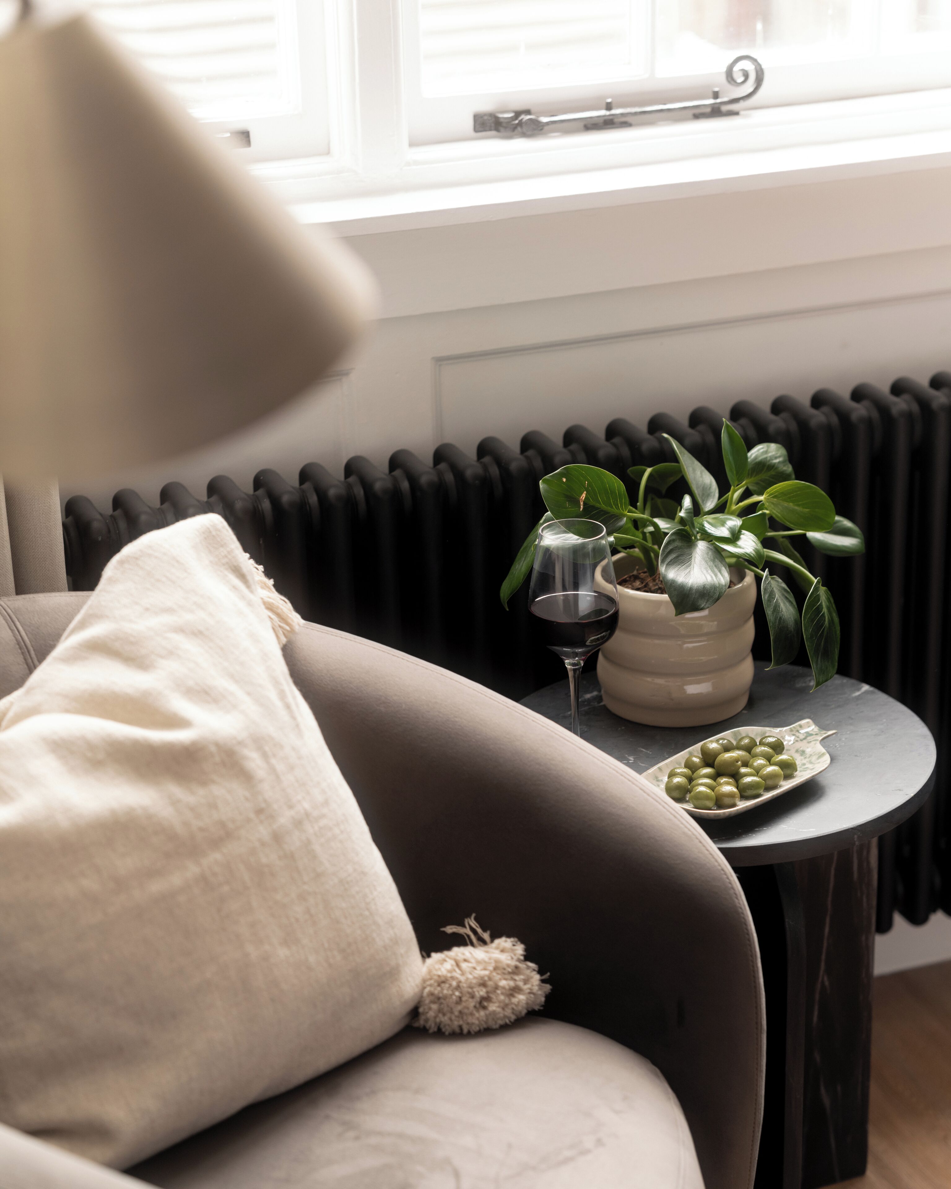 Un salon moderne avec un canapé gris, une petite table ronde ornée d’un pot de plantes, un verre à vin et une assiette d’olives vertes.