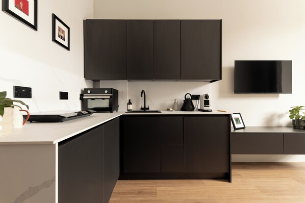 Une cuisine moderne avec des meubles de couleur sombre, un four encastré, une machine à café et un téléviseur à écran plat fixé au mur.