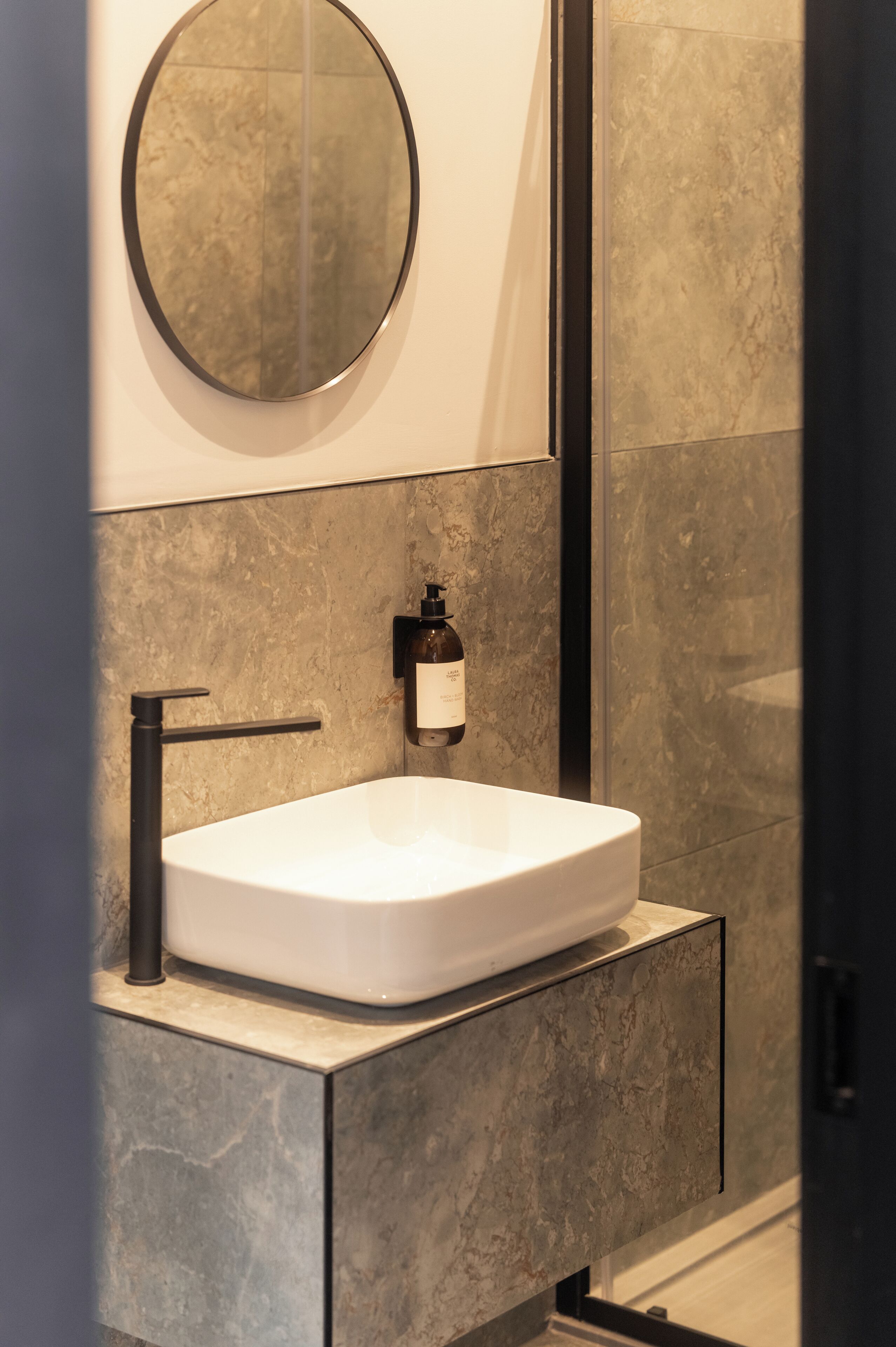 Un baño con un lavabo blanco, un espejo redondo y un dispensador de jabón en la pared.
