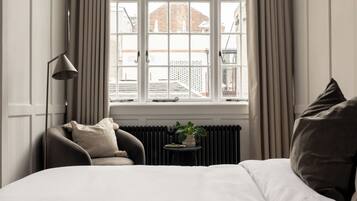 Une chambre d’hôtel avec un grand lit, une chaise, une petite table avec une plante et une fenêtre avec des rideaux.
