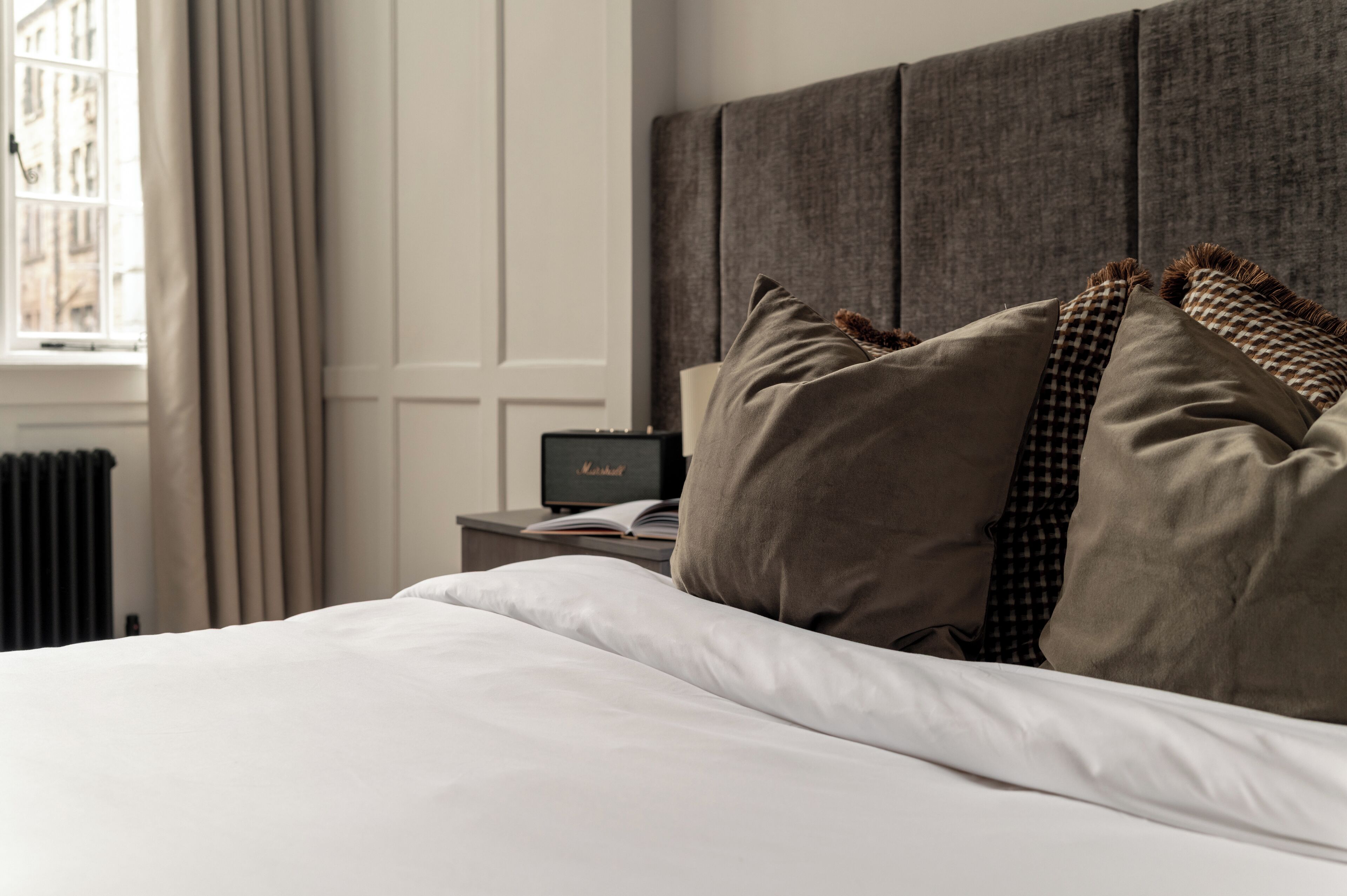 A neatly made bed with white linens and several pillows, a bedside table with a lamp and a book, and a window with curtains.