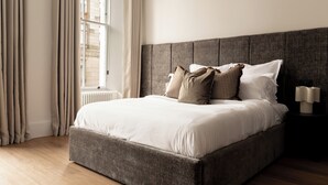 A neatly made bed with a grey headboard, beige curtains, and a window with blinds. - Moment Blythswood Street (Glasgow)