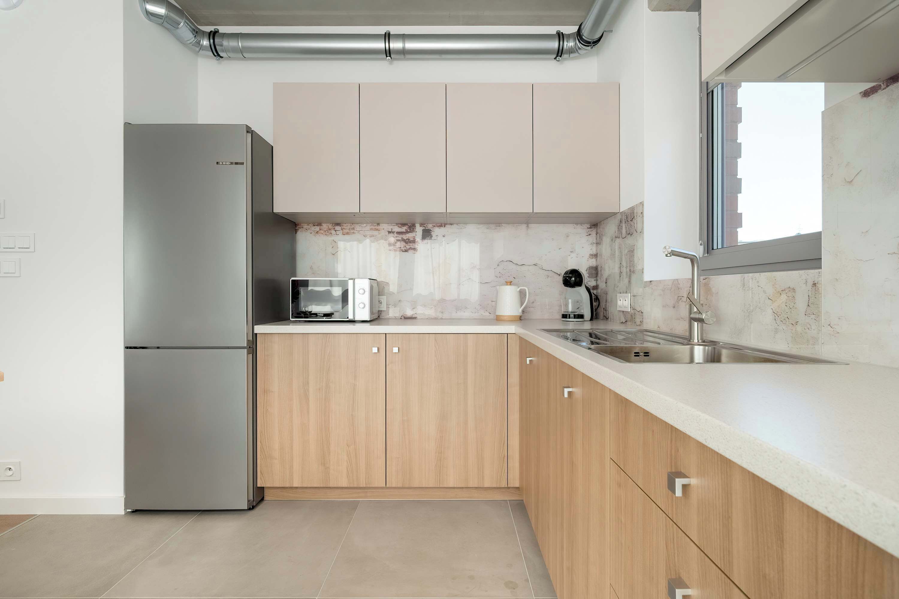 Una cocina moderna con un refrigerador de acero inoxidable, armarios de madera, una encimera blanca y un ventanal.