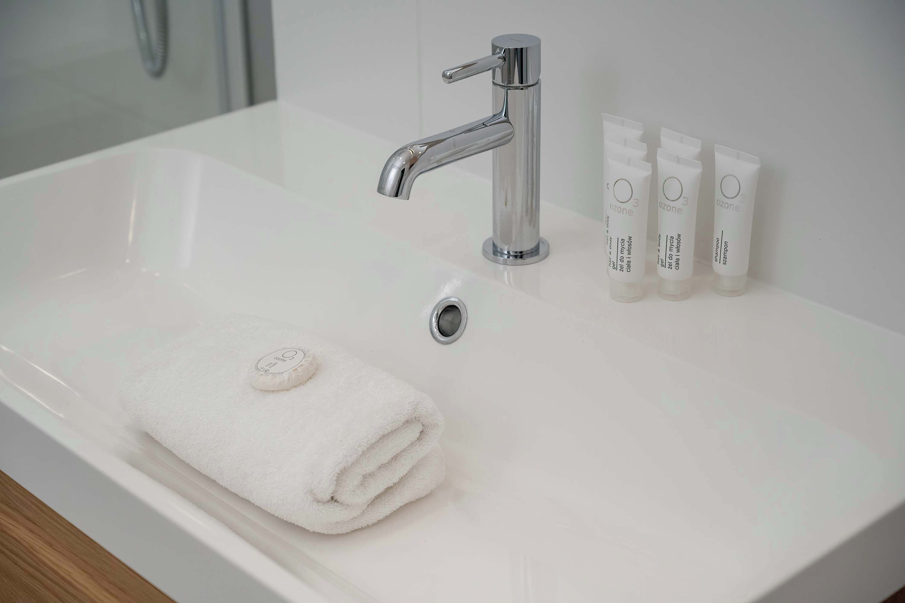 A white bathroom sink with a chrome faucet, a rolled white towel, and three Ozone shampoo bottles.