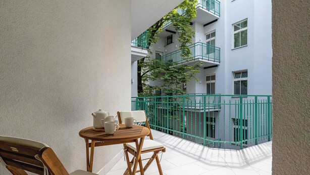 Een balkon met een houten tafel en stoelen, een kleine tafel met een theepot en kopjes, en een groene balustrade met uitzicht op een gebouw met meerdere ramen en balkons.