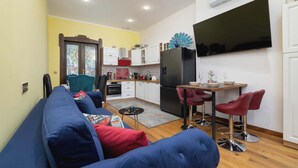 Een compacte leefruimte met een bank, een eettafel met twee stoelen, een keuken met kastjes en een koelkast, en een televisie aan de muur.