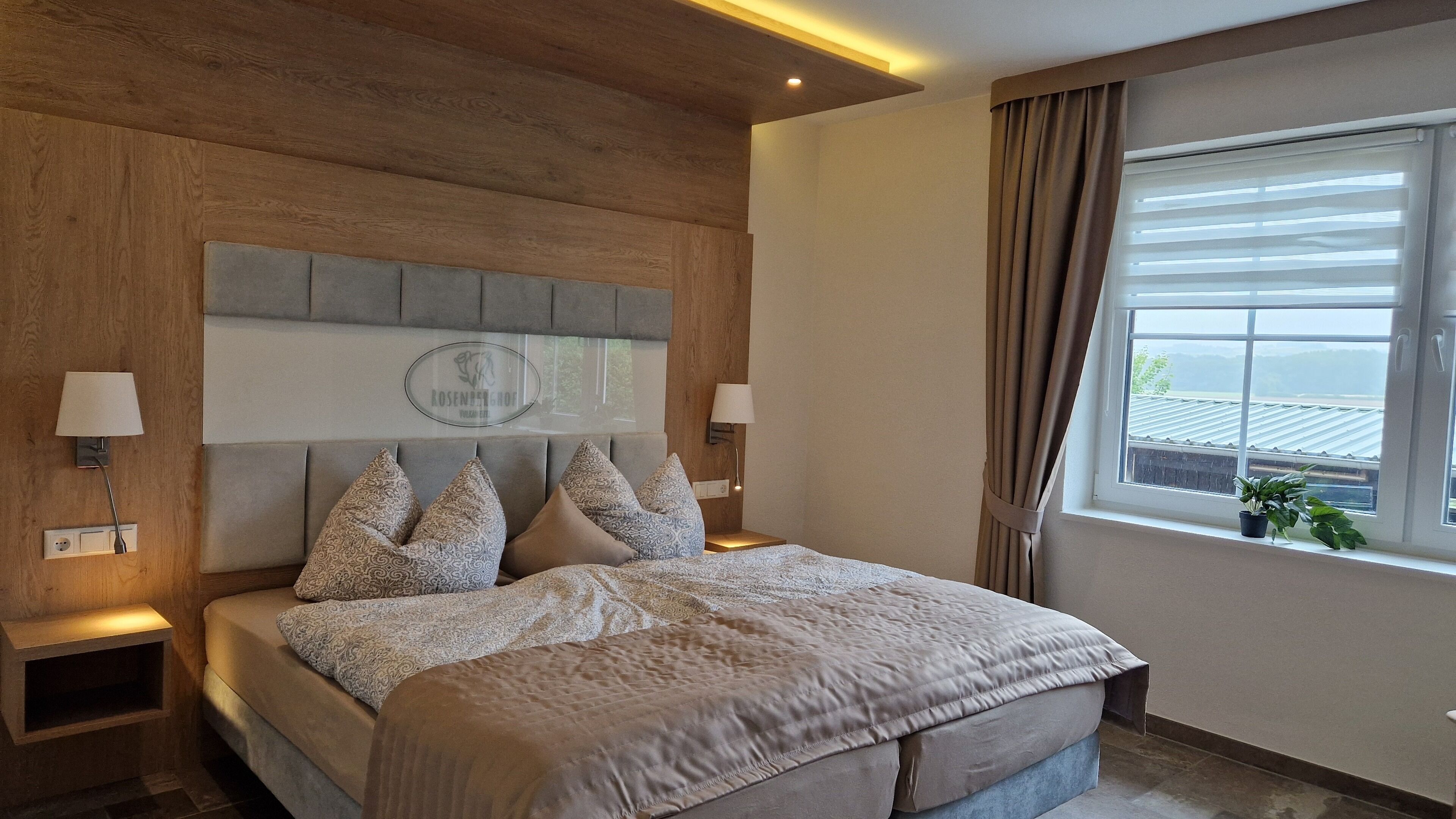 A modern bedroom with a wooden headboard, patterned bedding, bedside lamps, and a window with curtains.