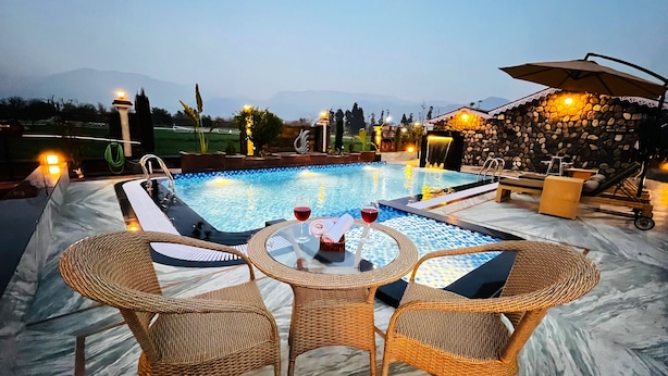 A swimming pool with a view of mountains, a table with chairs, and a stone wall with a water feature. - Vanhills : Boutique Stay in Jim Corbett (Kotabagh)