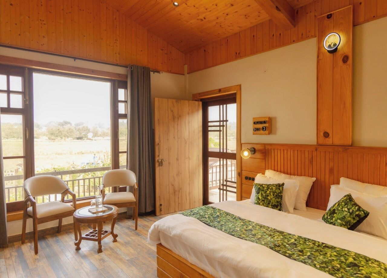 Een kamer met houten lambrisering, een bed, twee stoelen, een tafeltje en een groot raam.
