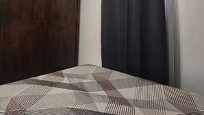 Une chambre avec une armoire en bois, un lit recouvert d’une couvre-lit à motifs, un rideau noir et un appareil de climatisation fixé au mur.