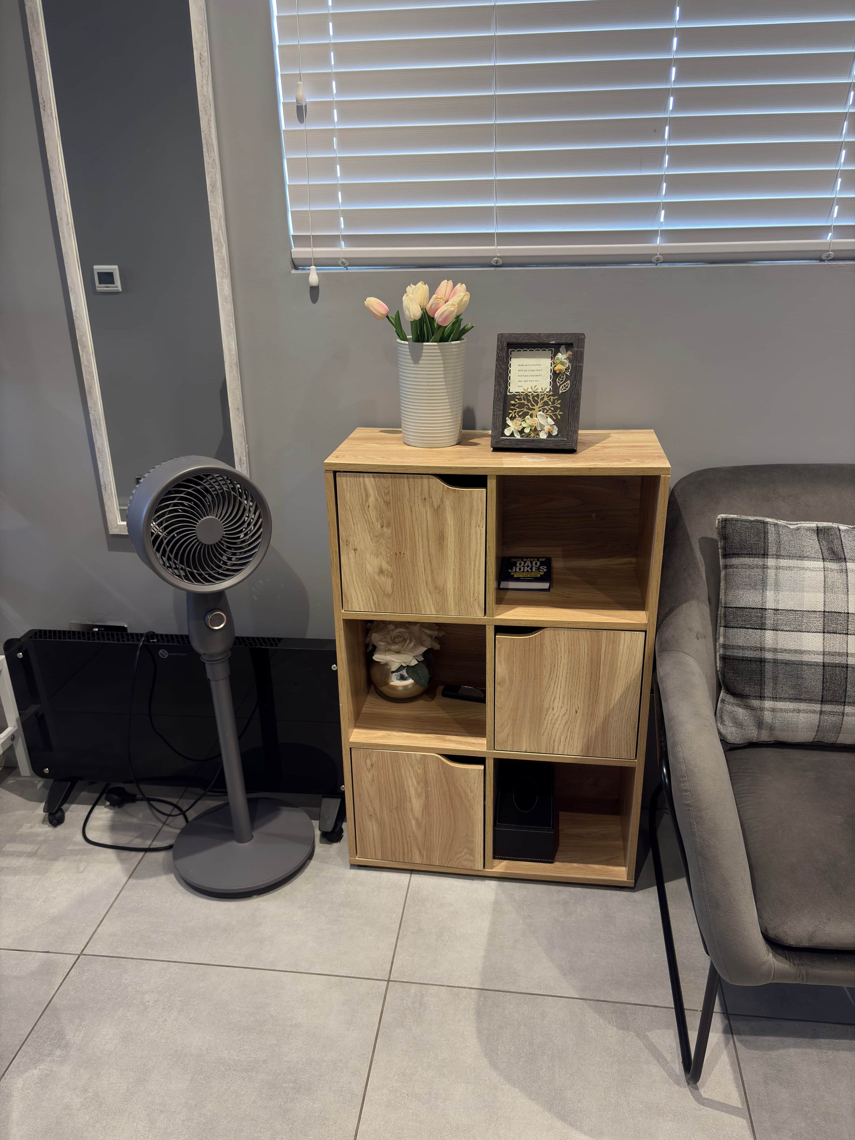 A room with a grey sofa, a wooden shelving unit with decorative items, a standing fan, and a radiator.