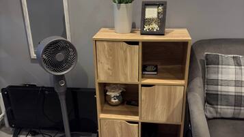 A room with a grey sofa, a wooden shelving unit with decorative items, a standing fan, and a radiator.