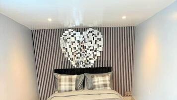 Een moderne slaapkamer met wanddecoratie in de vorm van een hart, een groot bed met grijze plaid, twee fauteuils en een gestreepte muur.