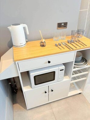 A kitchenette with a microwave, a kettle, and a wooden countertop with cutlery and glasses.