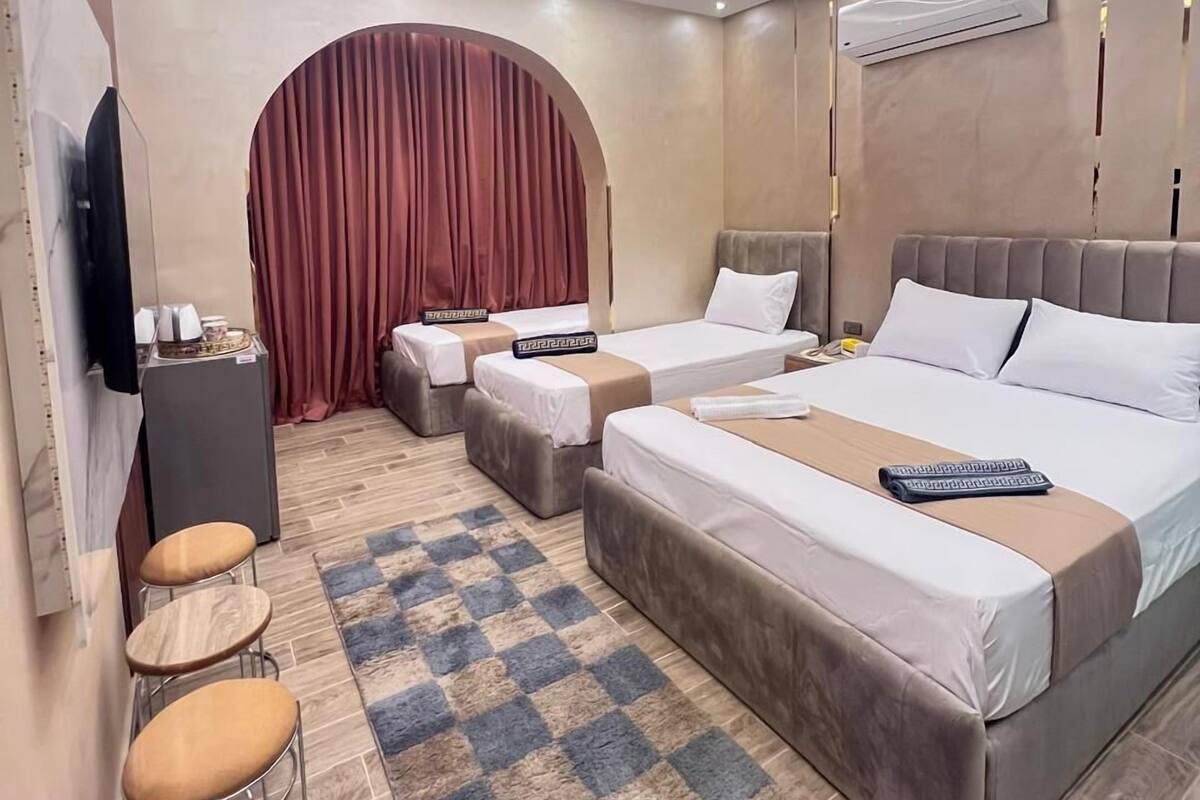 A modern hotel room with a large bed, two smaller beds, a TV, a minibar, a seating area with stools, and a checkered rug.
